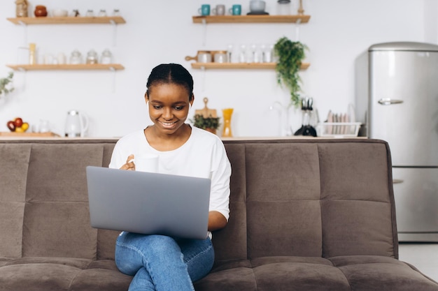 Joven afroamericana trabajando en una laptop sentada en un sofá en la cocina