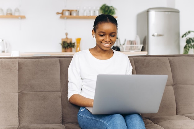 Joven afroamericana trabajando en una laptop sentada en un sofá en la cocina