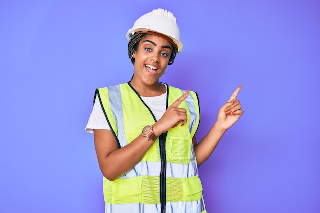 Joven afroamericana con trenzas con casco de seguridad y chaqueta reflectante sonriendo y mirando a la cámara apuntando con dos manos y dedos a un lado