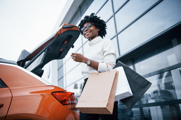 Joven afroamericana en vasos se encuentra al aire libre cerca del coche moderno después de comprar con paquetes.