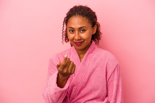 Joven afroamericana vistiendo una bata de baño aislada sobre fondo rosa apuntando con el dedo como si invitara a acercarse.