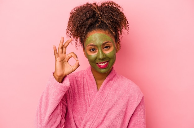 Joven afroamericana vistiendo una bata de baño y una máscara facial aislada sobre fondo rosa alegre y confiada mostrando un gesto bien.