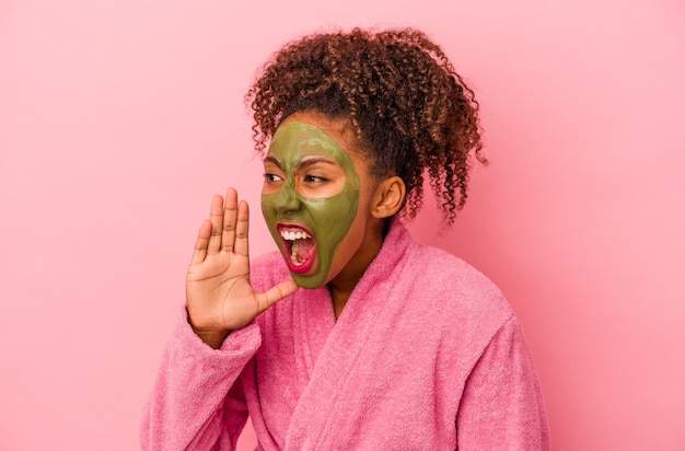 Joven afroamericana vistiendo una bata de baño y una máscara facial aislada sobre fondo rosa gritando y sosteniendo la palma cerca de la boca abierta.