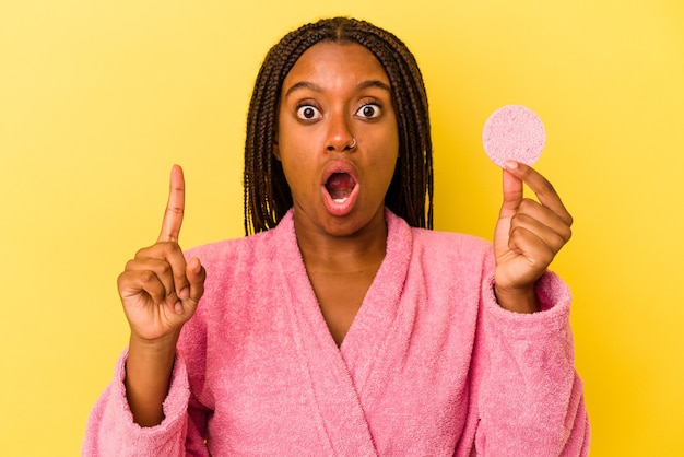 Joven afroamericana vistiendo una bata de baño sosteniendo un disco de maquillaje aislado sobre fondo amarillo con una gran idea, concepto de creatividad.