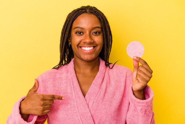 Joven afroamericana vistiendo una bata de baño sosteniendo un disco de maquillaje aislado sobre fondo amarillo persona apuntando con la mano a un espacio de copia de camisa, orgulloso y seguro