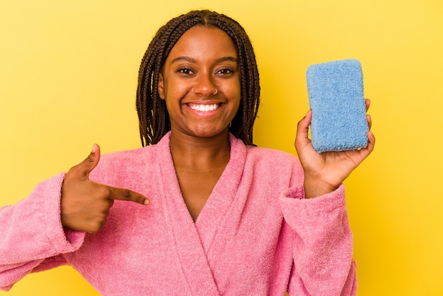 Joven afroamericana vistiendo una bata de baño sosteniendo una esponja azul aislada sobre fondo amarillo persona apuntando con la mano a un espacio de copia de camisa, orgulloso y seguro
