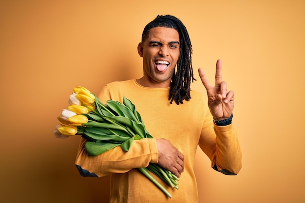 Joven afroamericano afro romántico con rastas sosteniendo ramo de tulipanes amarillos sonriendo con cara feliz guiñando un ojo a la cámara haciendo el signo de la victoria con los dedos Número dos