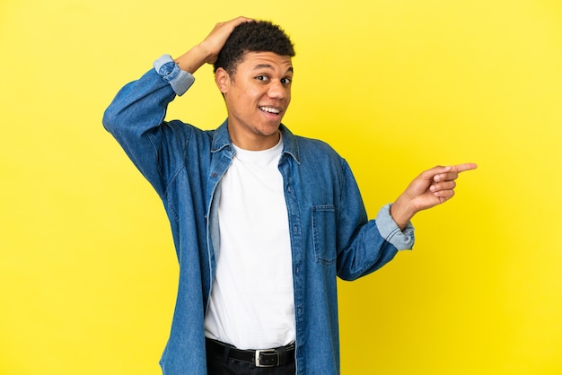 Joven afroamericano aislado sobre fondo amarillo sorprendido y apuntando con el dedo hacia el lado