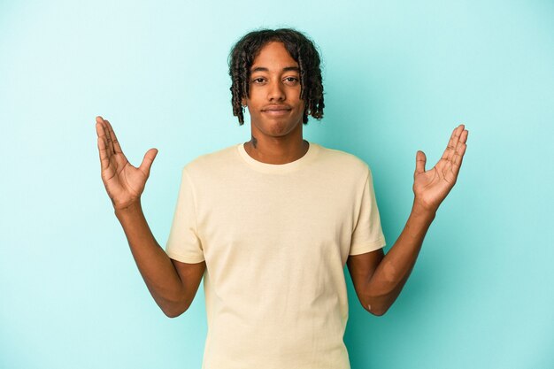 Joven afroamericano aislado sobre fondo azul sosteniendo algo poco con los dedos índices, sonriendo y confiado.