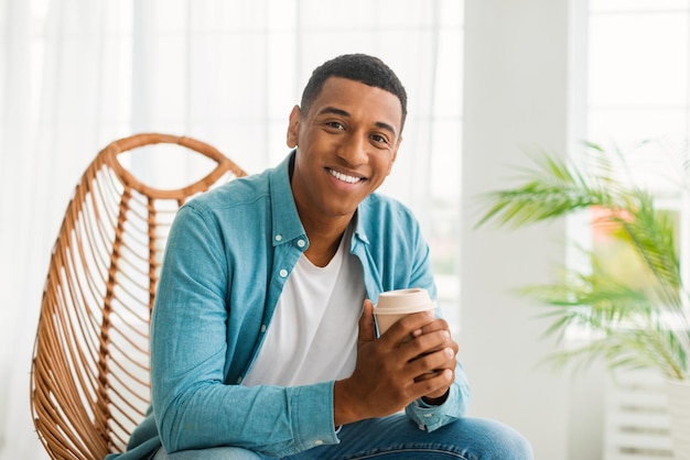 Un joven afroamericano alegre con una taza de café para llevar disfruta de la bebida y la calma en la sala de estar luminosa