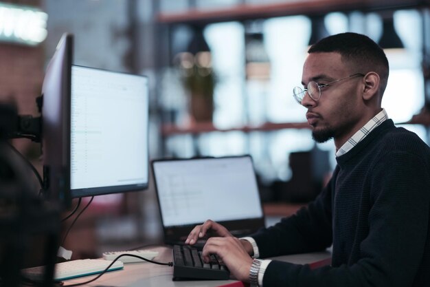 Joven afroamericano con anteojos sentado en una moderna oficina de coworking y trabajando en una computadora