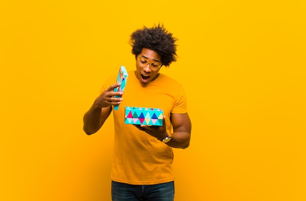 Joven afroamericano con una caja de regalo contra backgr naranja