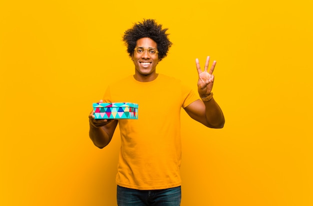 Joven afroamericano con una caja de regalo contra la pared naranja