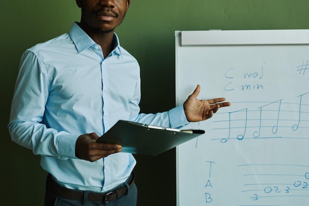 Joven afroamericano con camisa azul de pie junto a la pizarra con notas musicales y explicando th