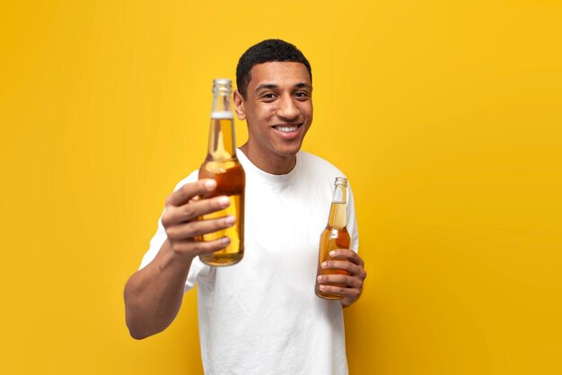 Joven afroamericano con camiseta blanca sostiene dos botellas de cerveza sobre fondo amarillo aislado