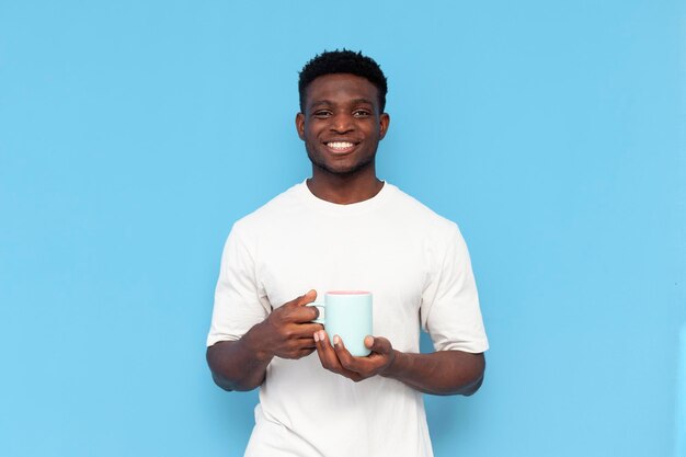 Un joven afroamericano con camiseta blanca sostiene una taza de café sobre fondo azul aislado