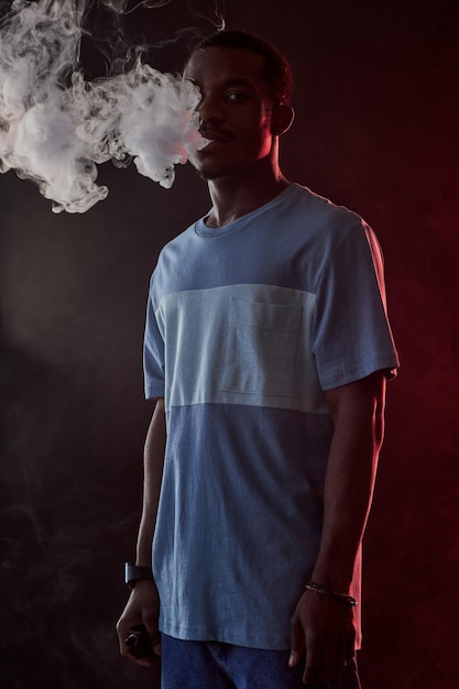 Joven afroamericano con camiseta y jeans fumando en la oscuridad