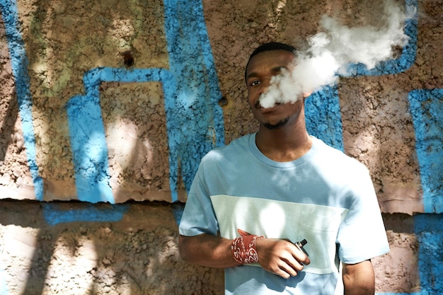 Joven afroamericano en camiseta de pie junto a la pared del edificio y fumando