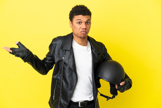 Joven afroamericano con un casco de motocicleta aislado sobre fondo amarillo con dudas mientras levanta las manos