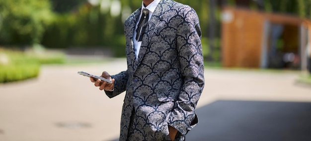 joven afroamericano con chaqueta y corbata negra en una calle sostiene un teléfono móvil en sus manos