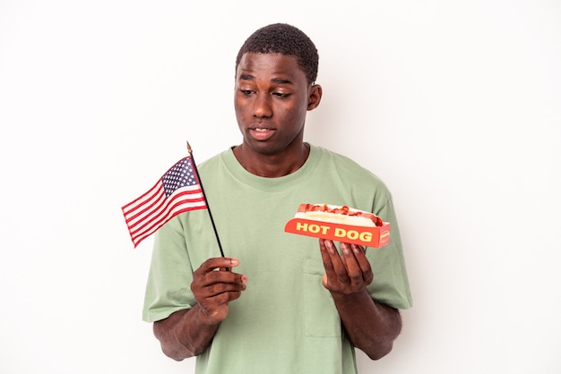Joven afroamericano comiendo hot dogs y sosteniendo la bandera americana aislado sobre fondo blanco.