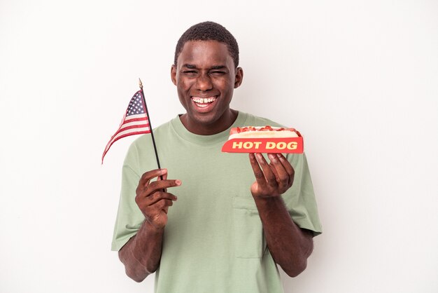 Joven afroamericano comiendo hot dogs y sosteniendo la bandera americana aislado sobre fondo blanco.