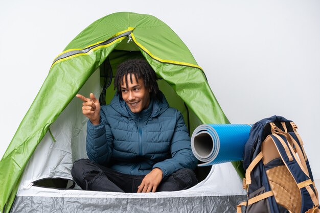 Joven afroamericano dentro de una tienda de campaña verde apuntando con el dedo hacia un lado y presentando un producto