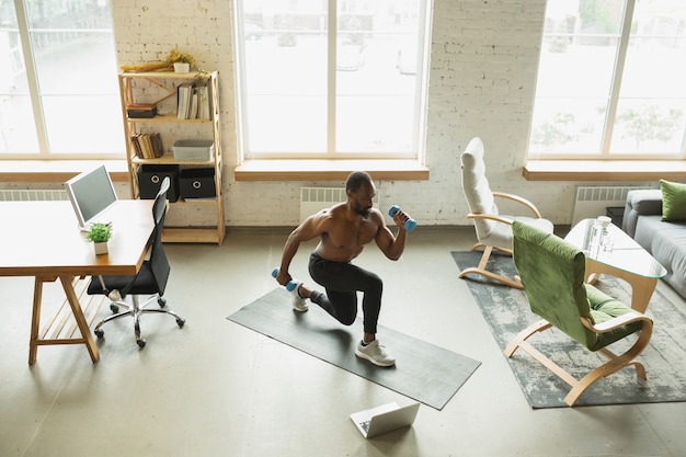 Joven afroamericano entrenando en casa durante la cuarentena del brote de coronavirus, haciendo ejercicios de fitness, aeróbicos. Mantenerse deportivo durante el aislamiento. Bienestar, concepto de movimiento. Abdominales.