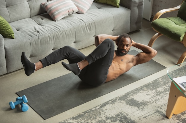 Joven afroamericano entrenando en casa durante la cuarentena del brote de coronavirus, haciendo ejercicios de fitness, aeróbicos. Mantenerse deportivo durante el aislamiento. Bienestar, concepto de movimiento. Ejercicio para los abdominales.