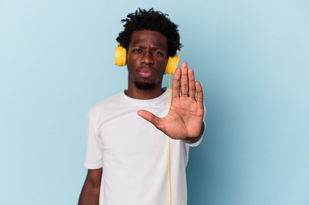 Joven afroamericano escuchando música aislada sobre fondo azul de pie con la mano extendida mostrando la señal de stop, impidiéndote.