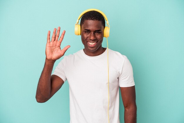 Joven afroamericano escuchando música aislado de fondo azul sonriendo alegre mostrando el número cinco con los dedos.