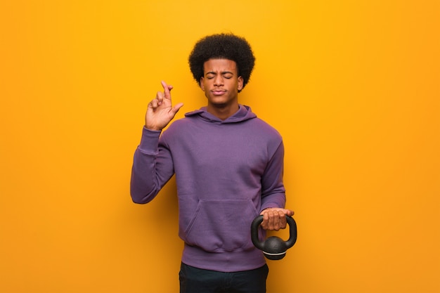 Joven afroamericano fitness hombre sosteniendo una pesa cruzando los dedos para tener suerte