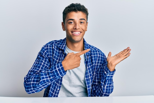 Foto joven afroamericano guapo con ropa informal sentado en la mesa asombrado y sonriendo a la cámara mientras presenta con la mano y señala con el dedo.