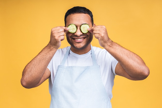 Joven afroamericano indio sosteniendo rodajas de pepino sobre los ojos y sonreír o reír. Aislado sobre fondo amarillo. Cocine preparando la comida.