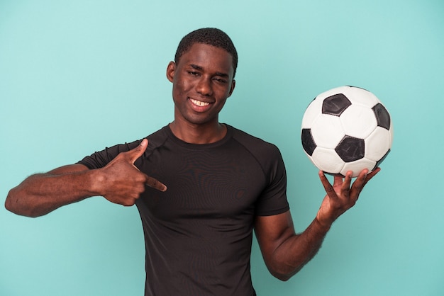 Joven afroamericano jugando al fútbol aislado sobre fondo azul persona apuntando con la mano a un espacio de copia de camisa, orgulloso y seguro