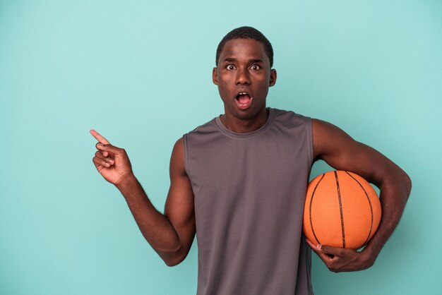Joven afroamericano jugando baloncesto aislado de fondo azul apuntando hacia un lado