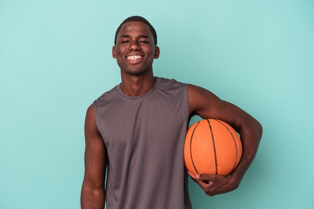 Joven afroamericano jugando baloncesto aislado de fondo azul feliz sonriendo y alegre