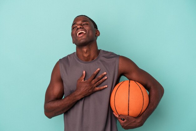 Un joven afroamericano jugando baloncesto aislado de fondo azul se ríe a carcajadas manteniendo la mano en el pecho.