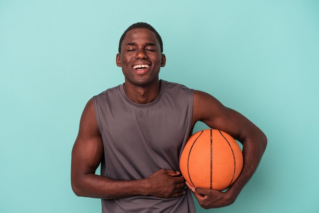 Joven afroamericano jugando baloncesto aislado de fondo azul riéndose y divirtiéndose.