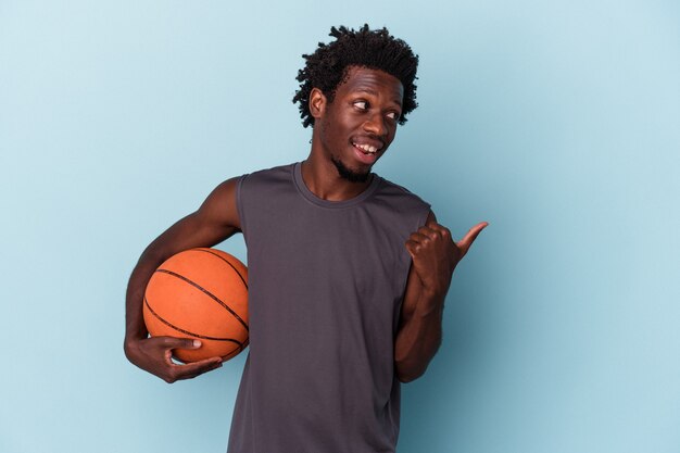 Joven afroamericano jugando baloncesto aislado sobre fondo azul apunta con el dedo pulgar, riendo y despreocupado.