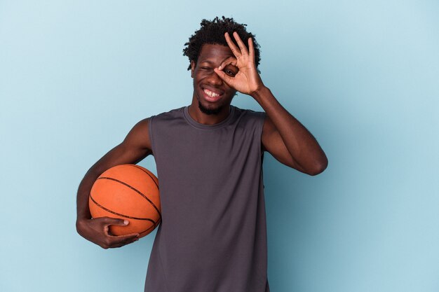 Joven afroamericano jugando baloncesto aislado sobre fondo azul emocionado manteniendo el gesto ok en el ojo.