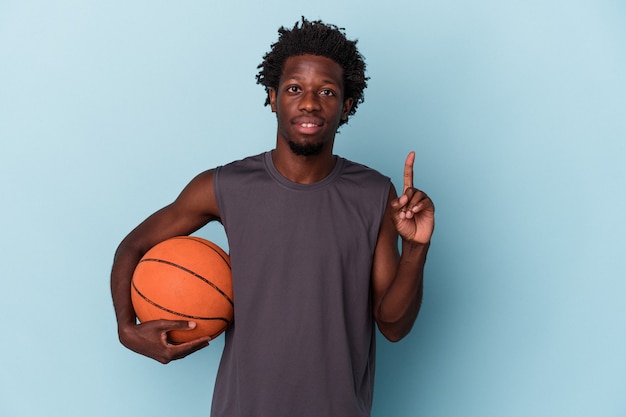 Joven afroamericano jugando baloncesto aislado sobre fondo azul mostrando el número uno con el dedo.