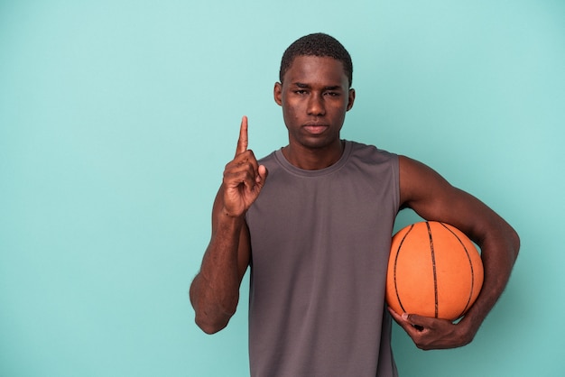 Joven afroamericano jugando baloncesto aislado sobre fondo azul mostrando el número uno con el dedo.