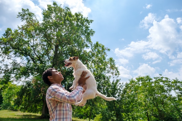 Joven afroamericano lanza perro Jack Russell terrier en el parque al aire libre en verano. Caminando en el parque en verano, vista de ángulo bajo