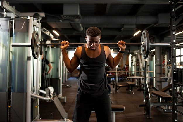 Un joven afroamericano levanta una barra en un gimnasio oscuro. Un deportista se entrena en una sala de fitness.