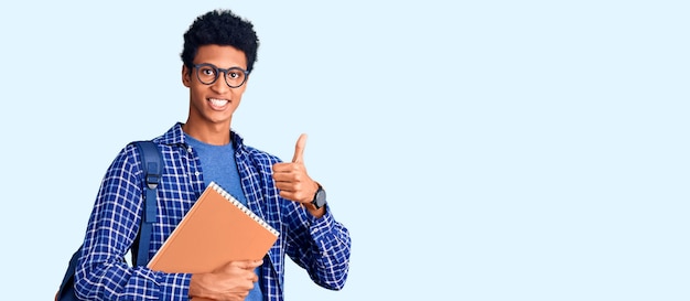 Joven afroamericano con mochila de estudiante sosteniendo un libro haciendo un gesto feliz con los pulgares hacia arriba con la mano. expresión de aprobación mirando a la cámara que muestra el éxito.