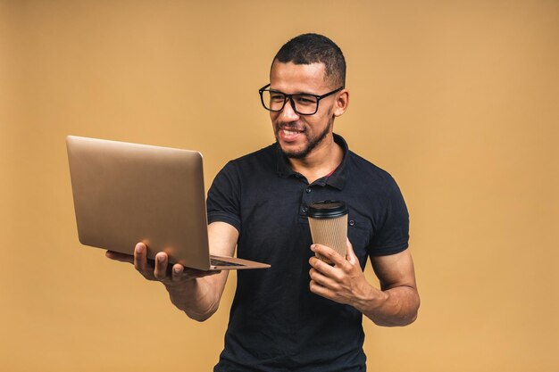 Joven afroamericano negro sonriente de pie y usando una computadora portátil aislada sobre fondo beige Bebiendo café