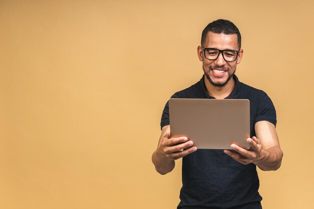 Joven afroamericano negro sonriente de pie y usando una computadora portátil aislada sobre un fondo beige