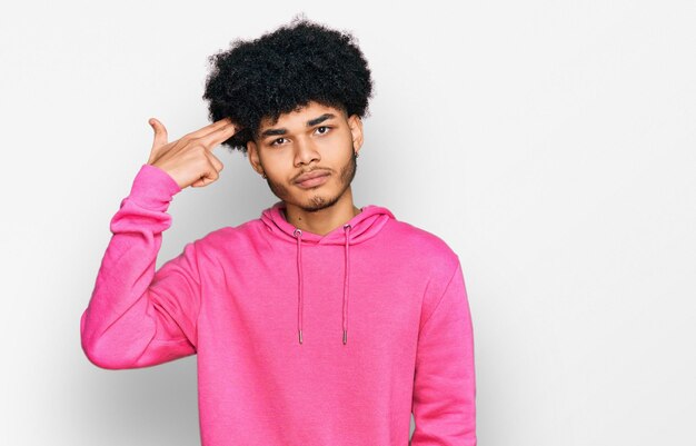 Joven afroamericano con el pelo afro usando una sudadera rosa casual disparándose y matándose apuntando con la mano y los dedos a la cabeza como un arma, gesto suicida.