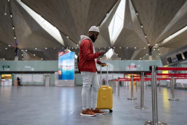 Joven afroamericano que usa un teléfono inteligente que viaja con equipaje dentro de la terminal del aeropuerto